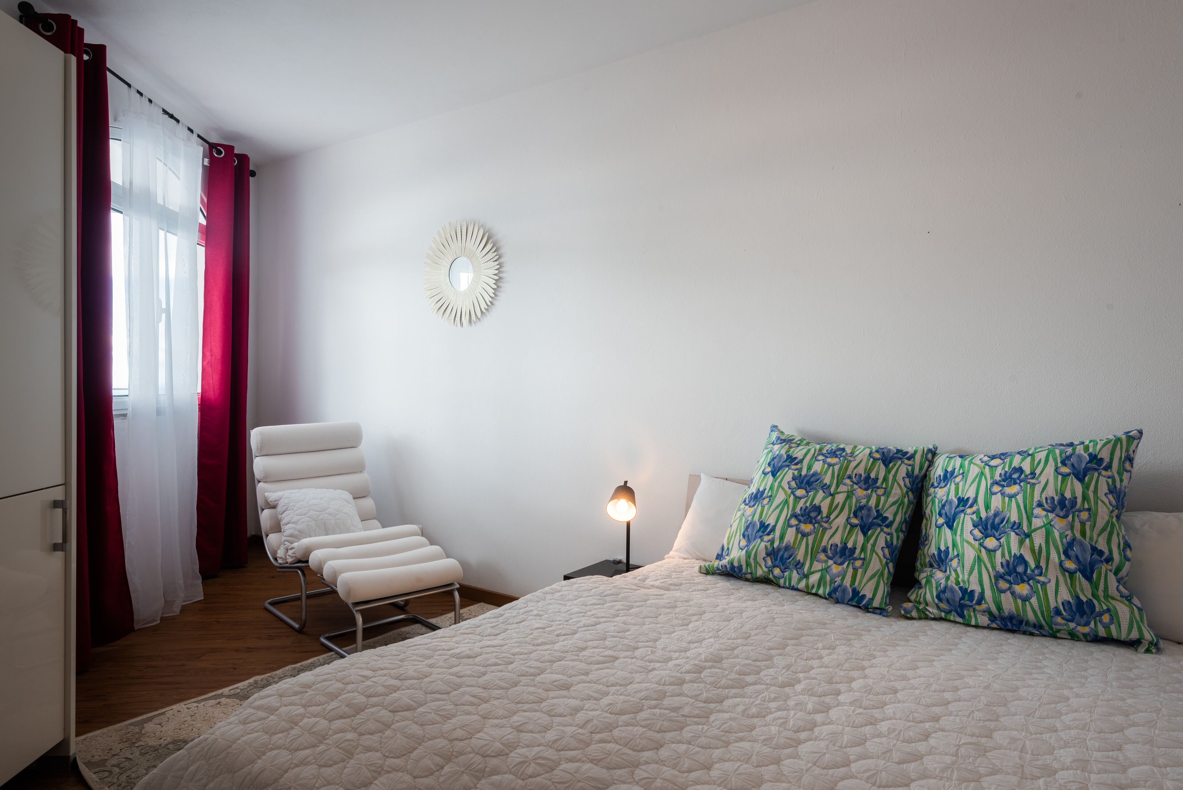 A bedroom with a bed, a chair, a nightstand, a lamp, and a wall-mounted mirror.