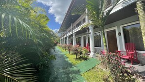 Un hotel con una galería techada, palmeras y sillas rojas.
