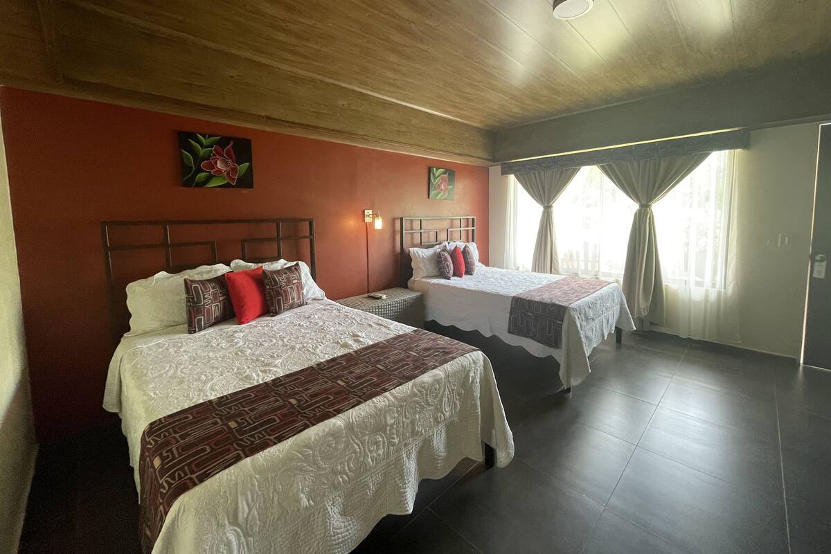 Ein Hotelzimmer mit zwei Betten, einer Holzbalkendecke, einer roten Wand und einem Fenster mit Vorhängen.
