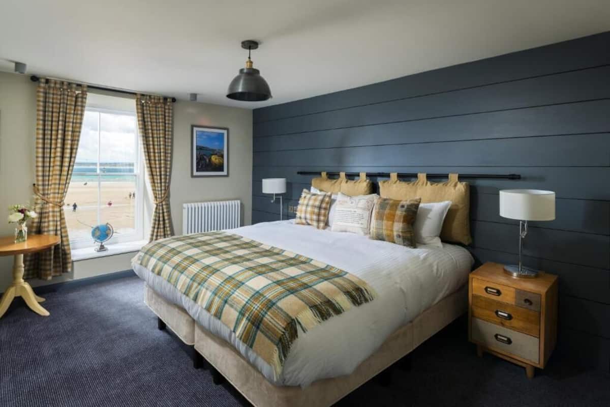 A bedroom with a bed, bedside tables, a window with curtains, a lamp, and a view of the beach.