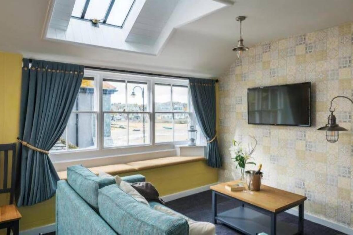 Ein Zimmer mit einer Couch, einem Tisch, einem Fernseher, einem Fenster mit Vorhängen und einem Dachfenster.