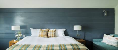 A hotel room with a bed, two bedside tables, a nightstand, a lamp, a chair, and a wall-mounted light fixture.