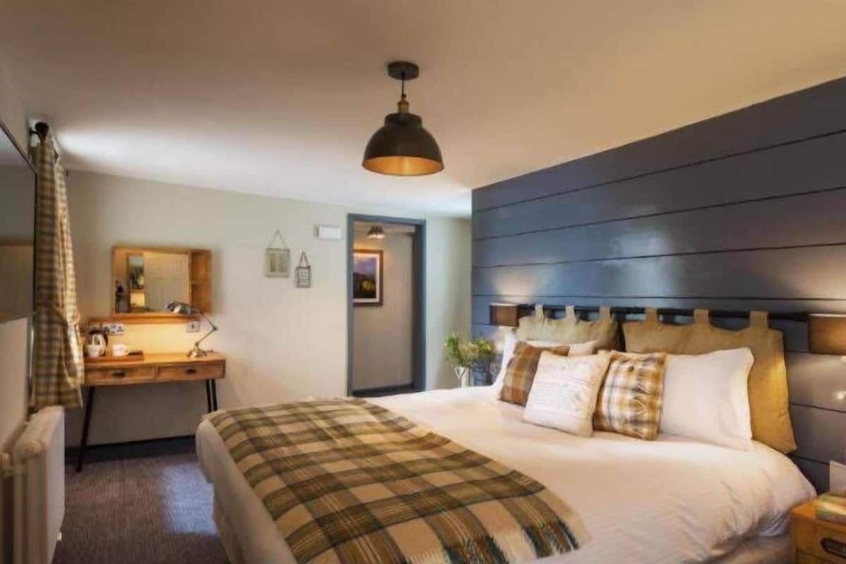 A hotel room with a bed, a desk, a mirror, a hanging light fixture, and a dark wooden headboard.