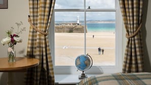 A room with a large window overlooking a beach, a globe on the windowsill, and a vase with flowers on a table. - Lifeboat Inn (St Ives)