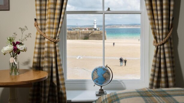 A room with a large window overlooking a beach, a globe on the windowsill, and a vase with flowers on a table. - Lifeboat Inn (St Ives)