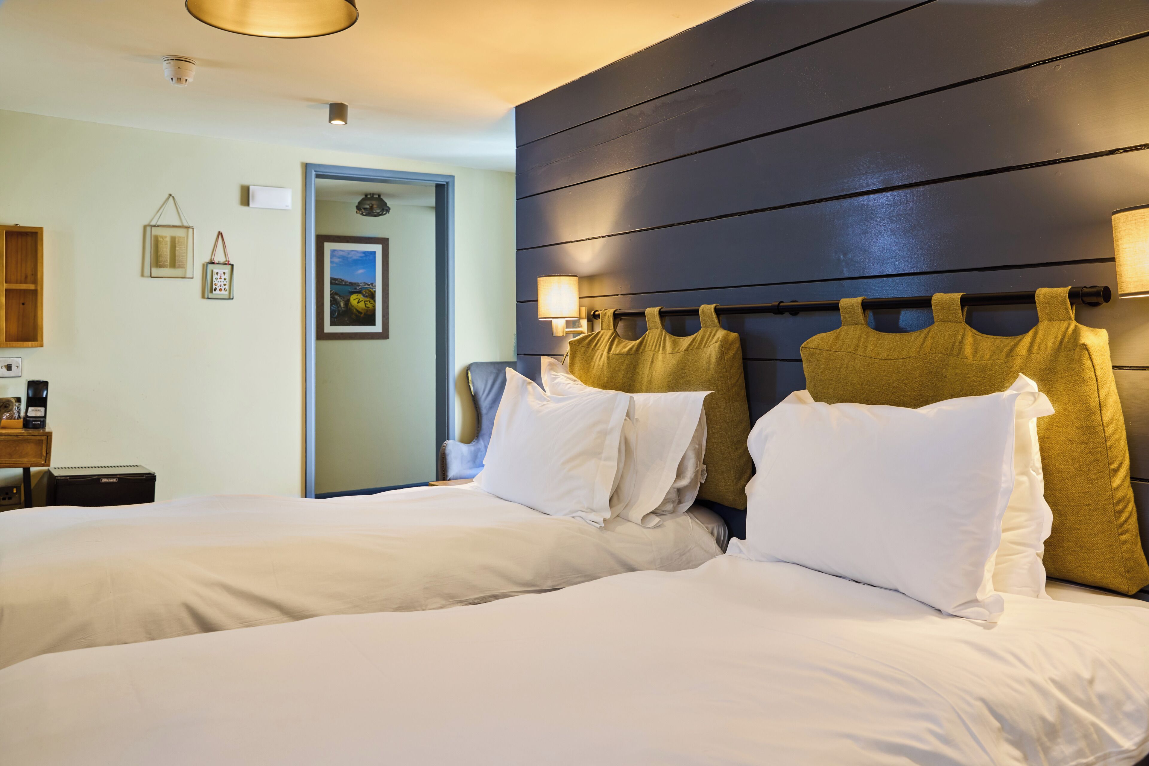 A hotel room with two beds, a bedside table, a lamp, and a door leading to another room.
