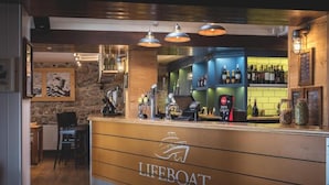 A bar area with a counter, bar stools, and a shelf with bottles. - Lifeboat Inn (St Ives)