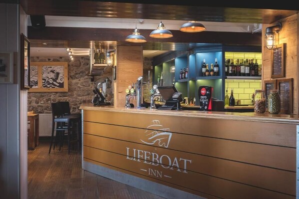 A bar area with a counter, bar stools, and a shelf with bottles. - Lifeboat Inn (St Ives)
