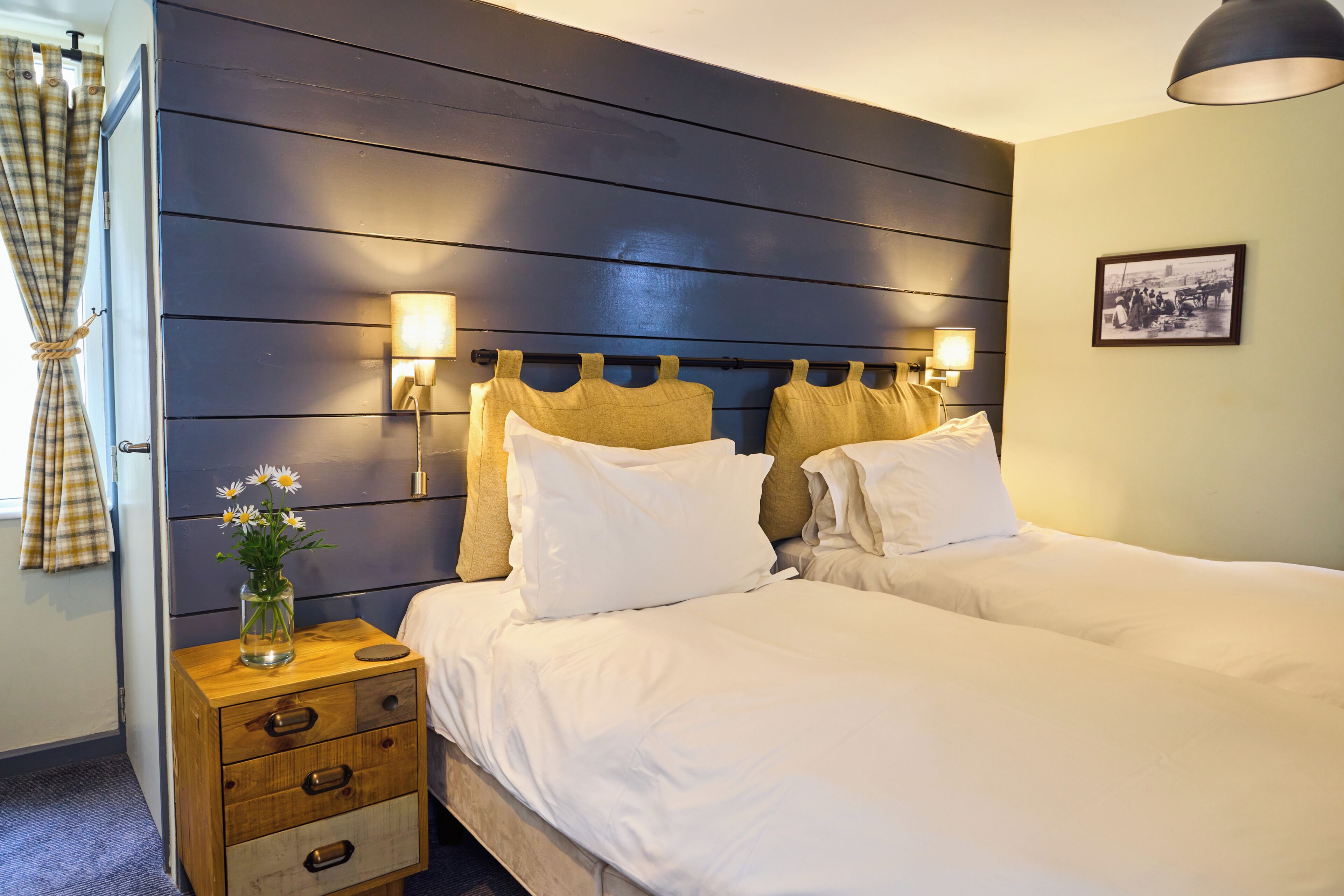A hotel room with a bed, bedside lamps, a nightstand with drawers, a vase with flowers, and a framed picture on the wall.