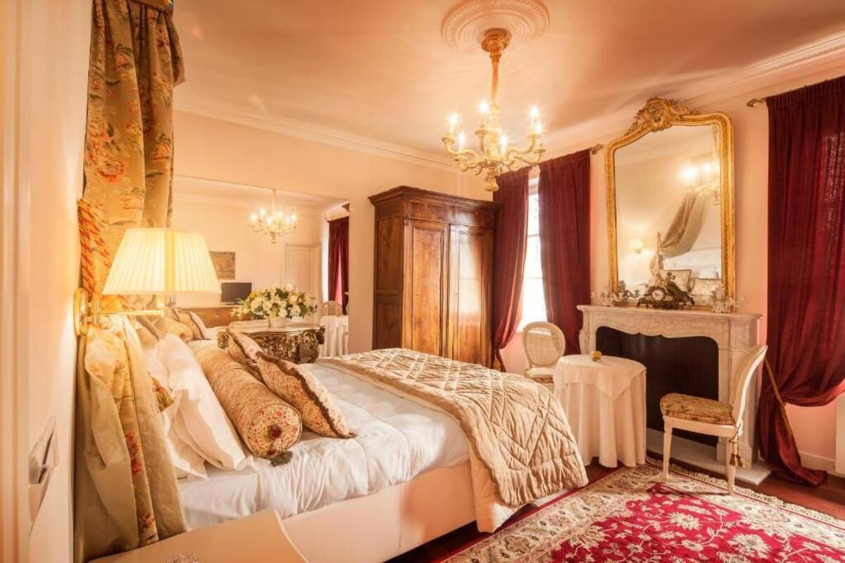 A classic bedroom with a large bed, a mirror, a fireplace, and a chandelier.