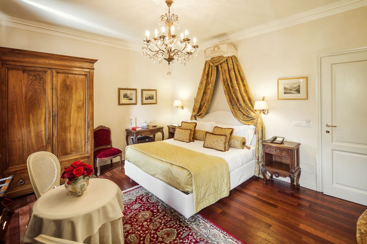 A well-appointed bedroom with a large bed, a chandelier, a wooden wardrobe, a red chair, and a small table with a vase of flowers.