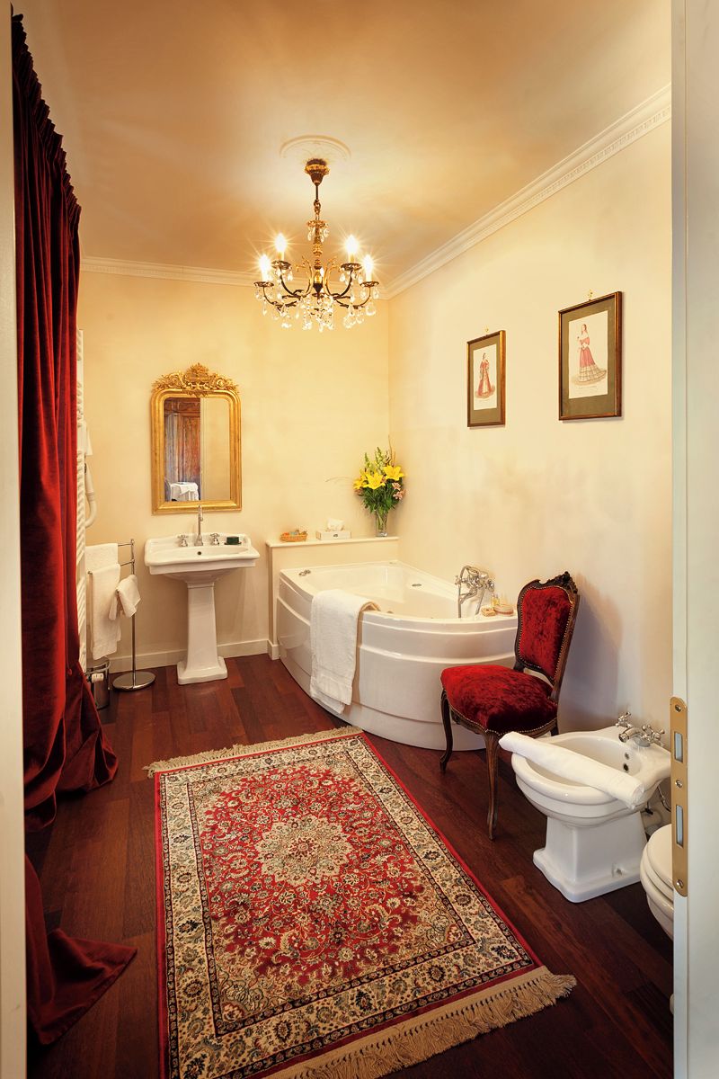 A bathroom with a pedestal sink, a freestanding bathtub, a red upholstered chair, a white toilet, and a chandelier.