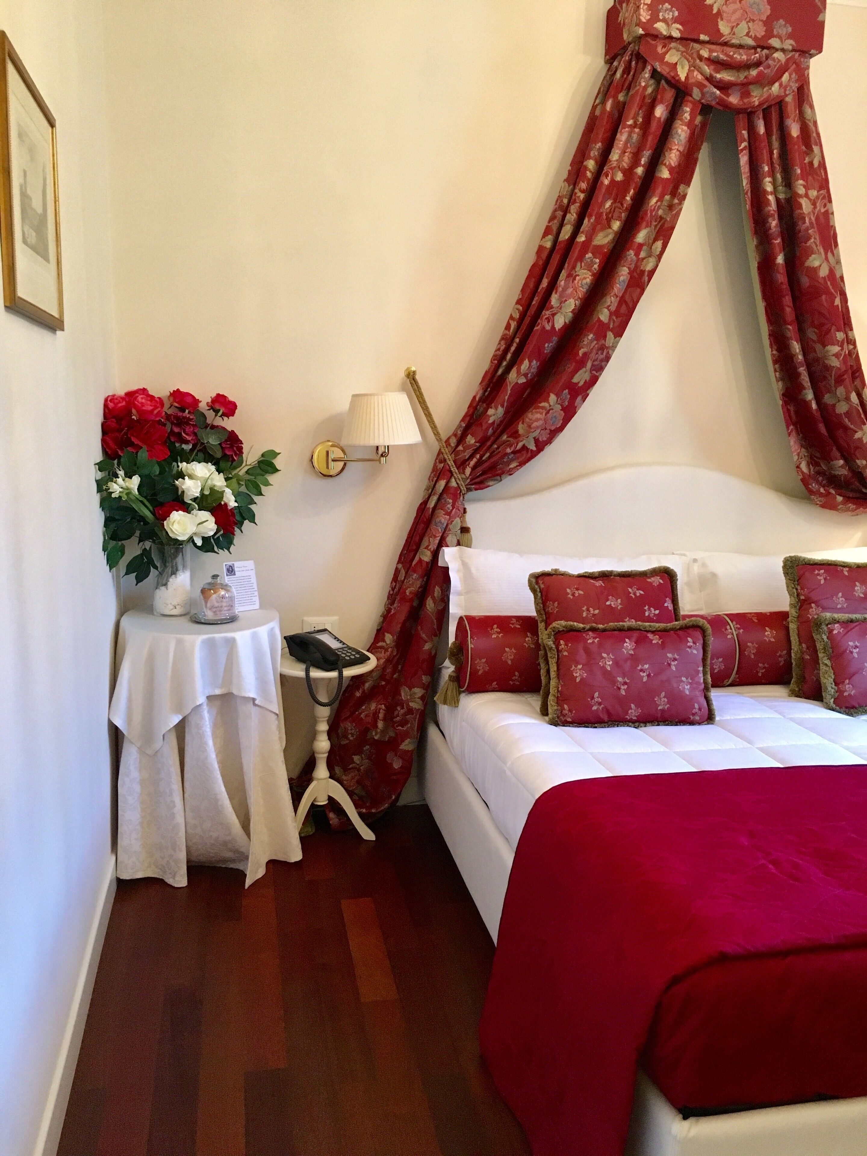 A hotel room with a bed, a bedside table with a lamp, a vase of flowers, and a red curtain.