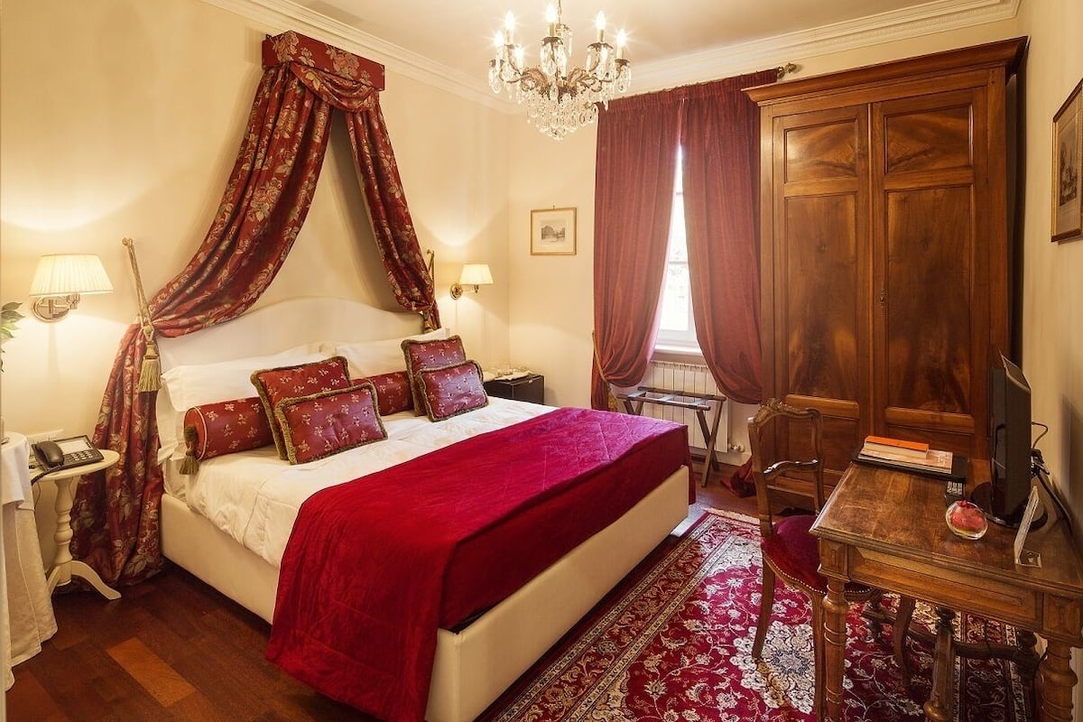 A bedroom with a large bed, a wooden wardrobe, a desk with a chair, and a chandelier.