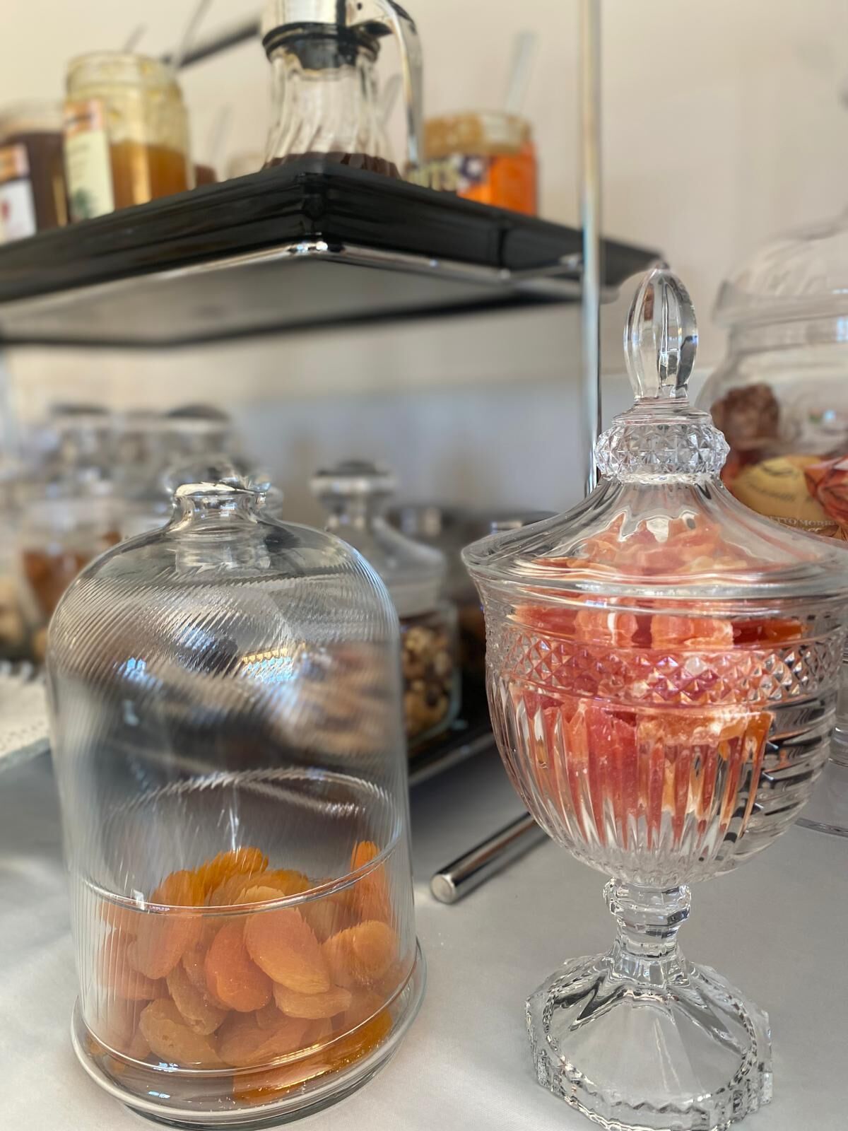 Disposição de buffet com recipiente de vidro coberto com frutas secas e uma tigela de vidro com tampa contendo uma fruta vermelha.