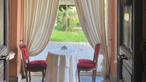 A room with two red upholstered chairs, a small round table, and a view of a garden through large curtains. - Duchessa Margherita (Vicoforte)