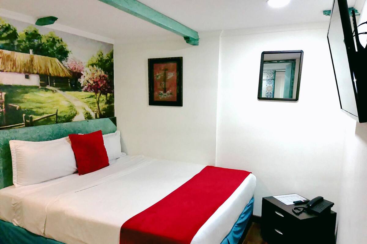 Un lit bien fait, recouvert d’une couverture rouge et blanche, une petite table de chevet avec un téléphone et une lampe, et un tableau représentant un paysage accroché au mur.