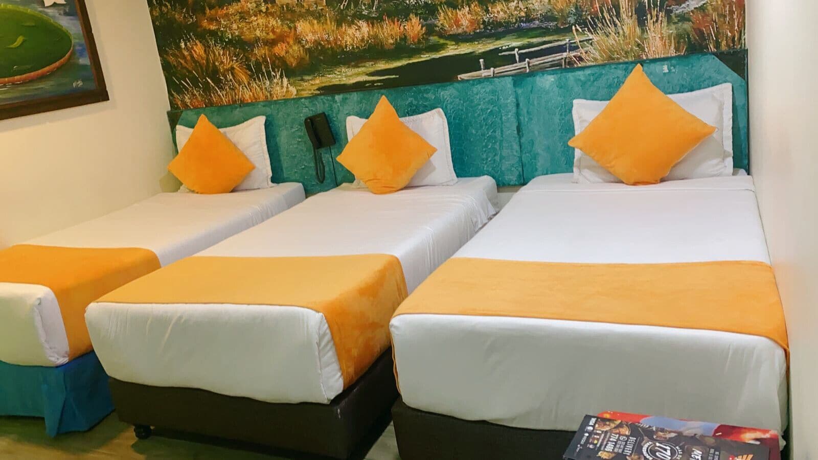 Habitación de hotel con tres camas individuales, cada una con colchón blanco y ropa de cama naranja. El cabecero es color verde azulado y tiene un teléfono instalado.