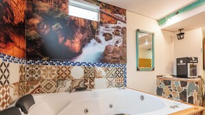 Een badkamer met een ruim bad, een spiegel, een koffieapparaat en een betegelde muur met een natuurdecoratie.