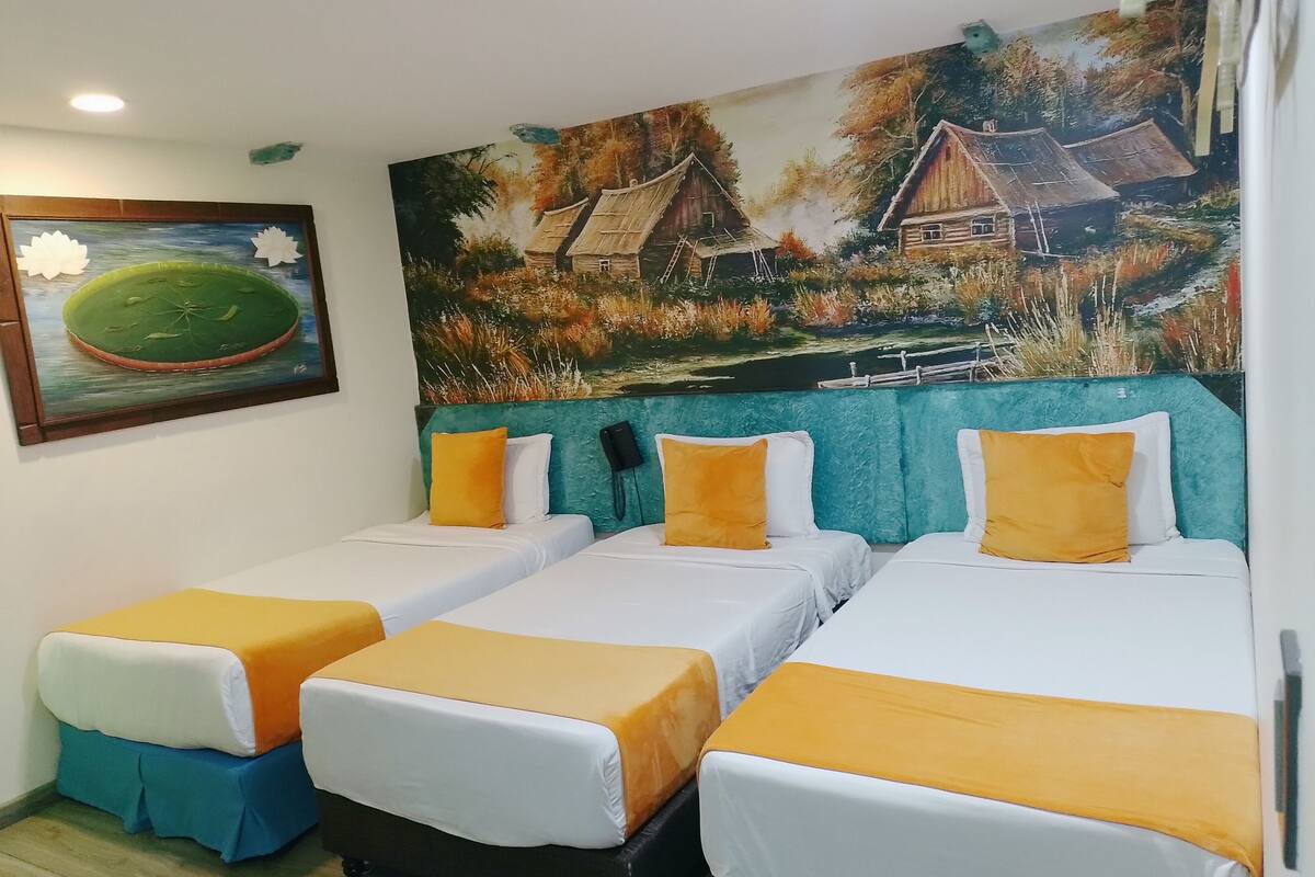 Une chambre d’hôtel avec deux lits simples, chacun recouvert d’une courtepointe blanche et orange, et un grand tableau représentant un paysage rural accroché au mur.