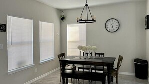 Un comedor con una mesa de madera oscura, sillas blancas y una araña. Hay tres jarrones blancos sobre la mesa, y un reloj en la pared.