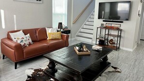 Un salón con un sofá de cuero marrón, una mesa de centro de madera oscura, un tapete de piel de vaca, un televisor montado en la pared y una escalera al fondo.