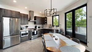 A modern kitchen with stainless steel appliances, a wooden dining table, and large windows offering a view of greenery.
