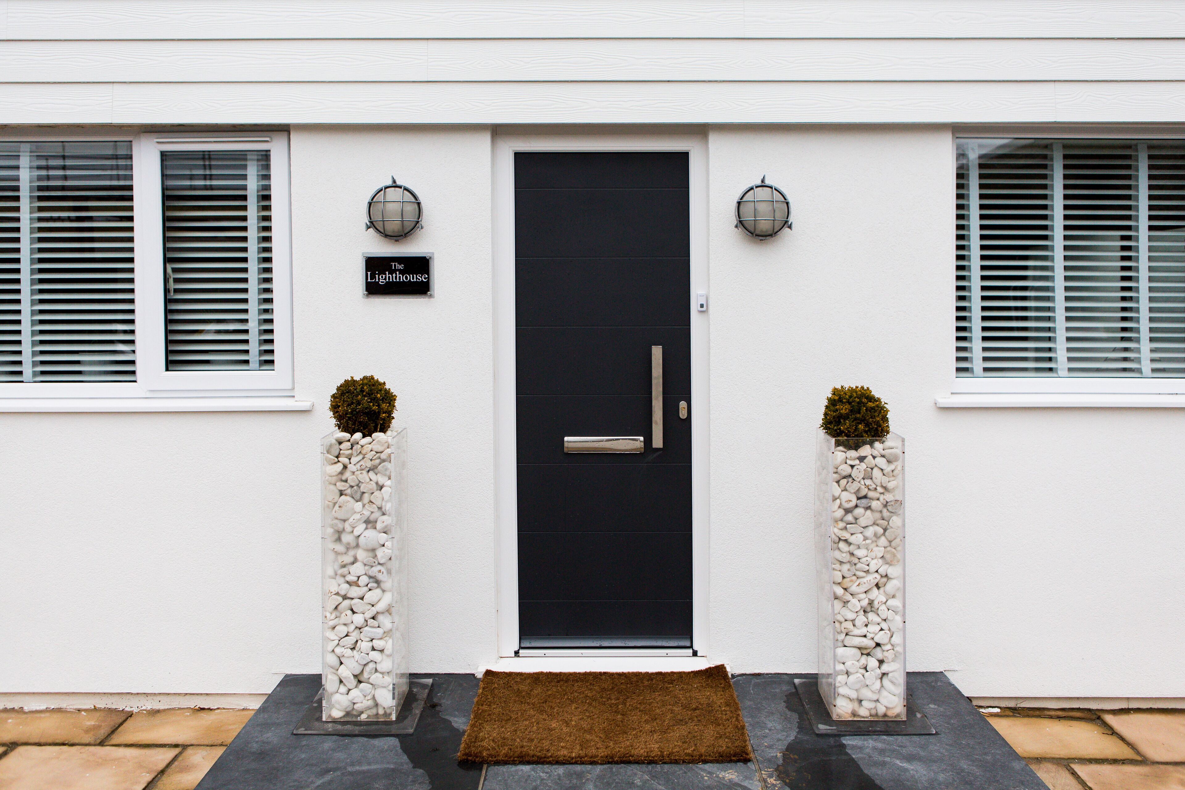 A modern house with a black front door, two potted plants, and a sign that reads "The Lighthouse."