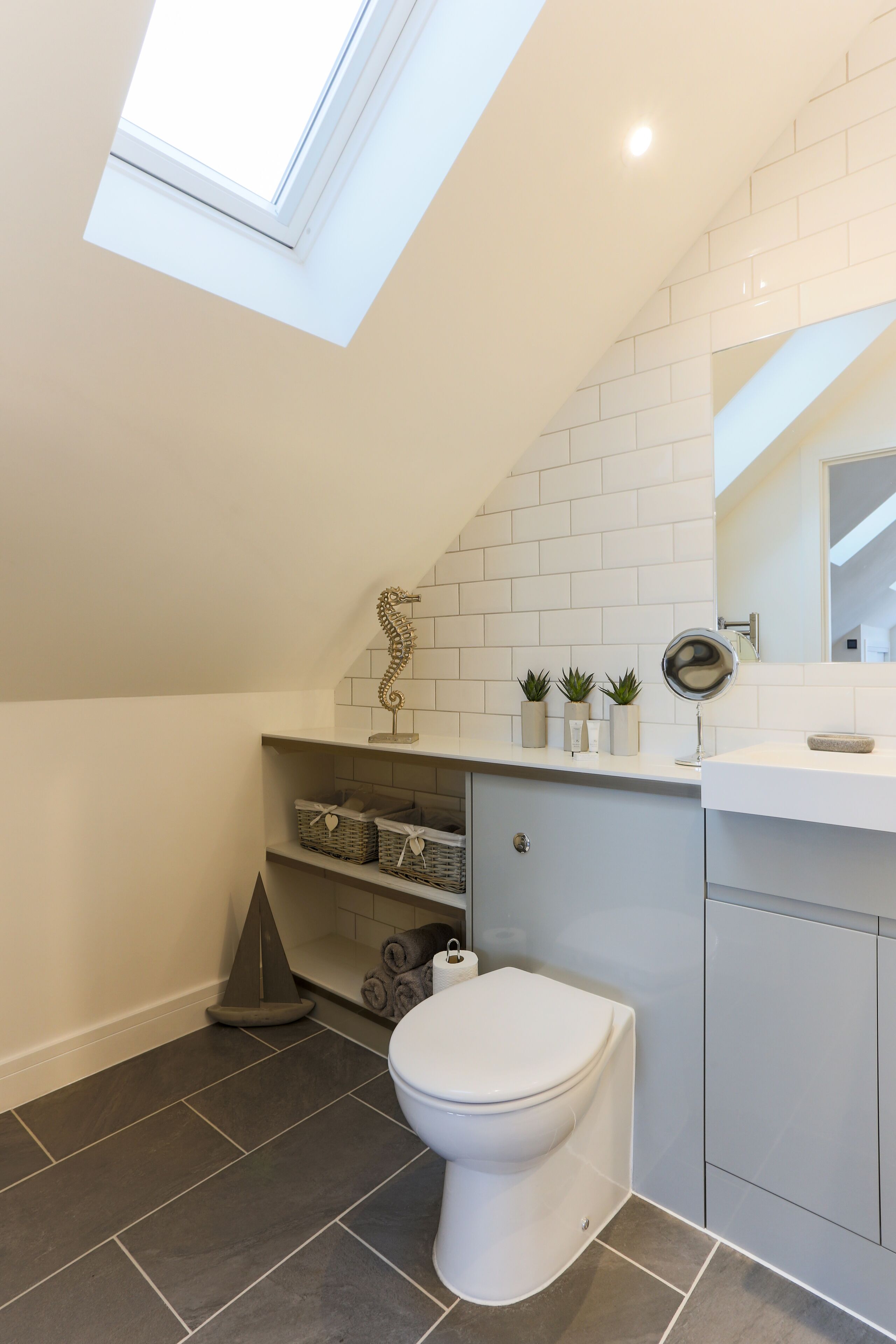 A modern bathroom with a skylight, white tiles, a toilet, a sink with a mirror, and decorative plants.