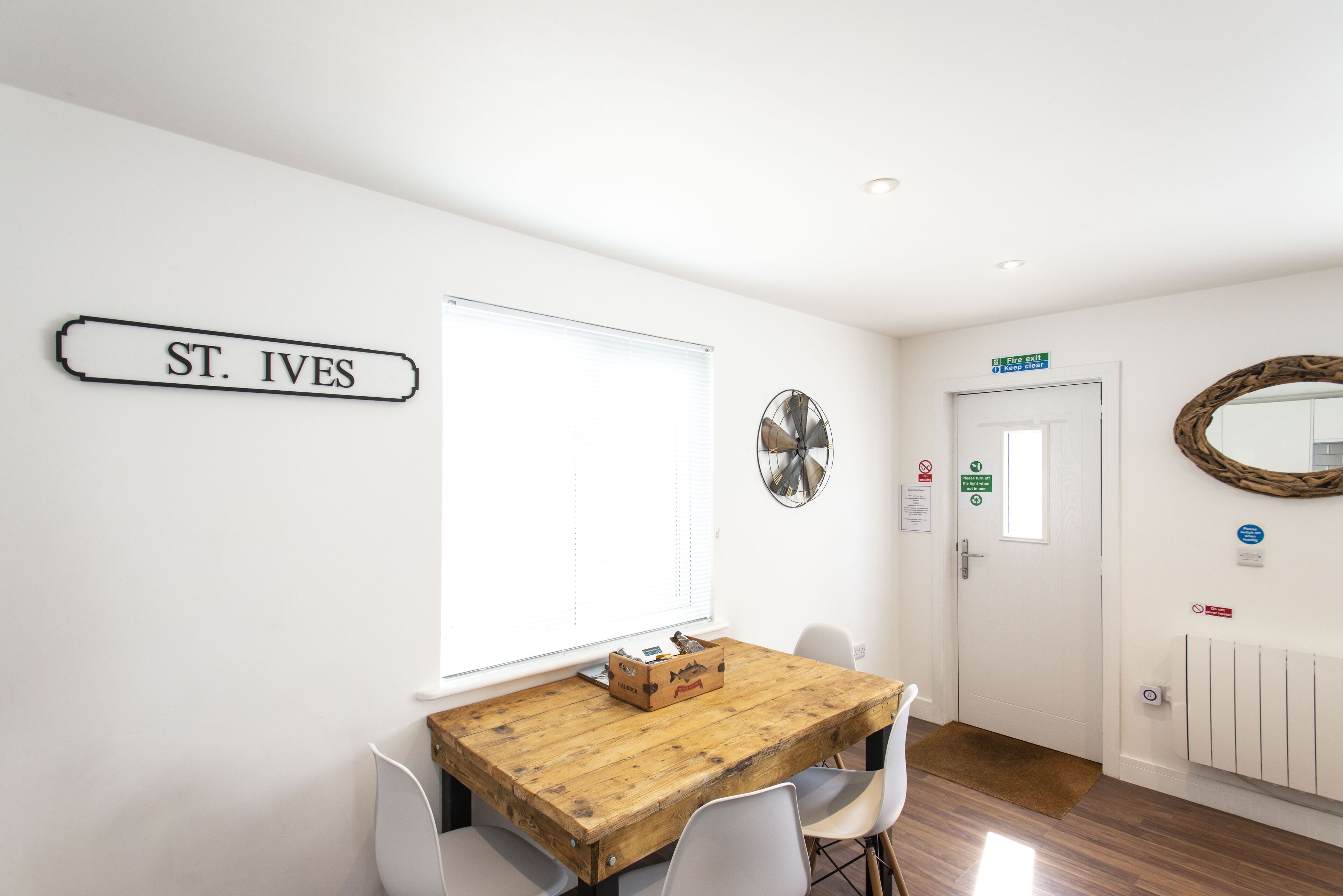 A room with a wooden table, white chairs, a sign reading "ST. IVES", a window with blinds, a door with a sign, and a wall-mounted fan.
