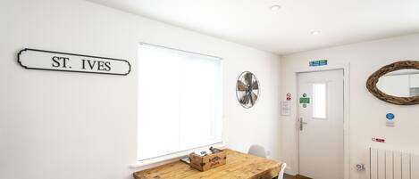 Una habitació amb taula de fusta, cadires blanques, un senyal que diu "ST. IVES", una finestra amb persianes, una porta amb senyal i un ventilador fixat a la paret.