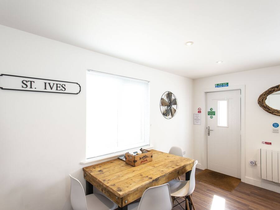A room with a wooden table, white chairs, a sign reading "ST. IVES", a window with blinds, a door with a sign, and a wall-mounted fan.