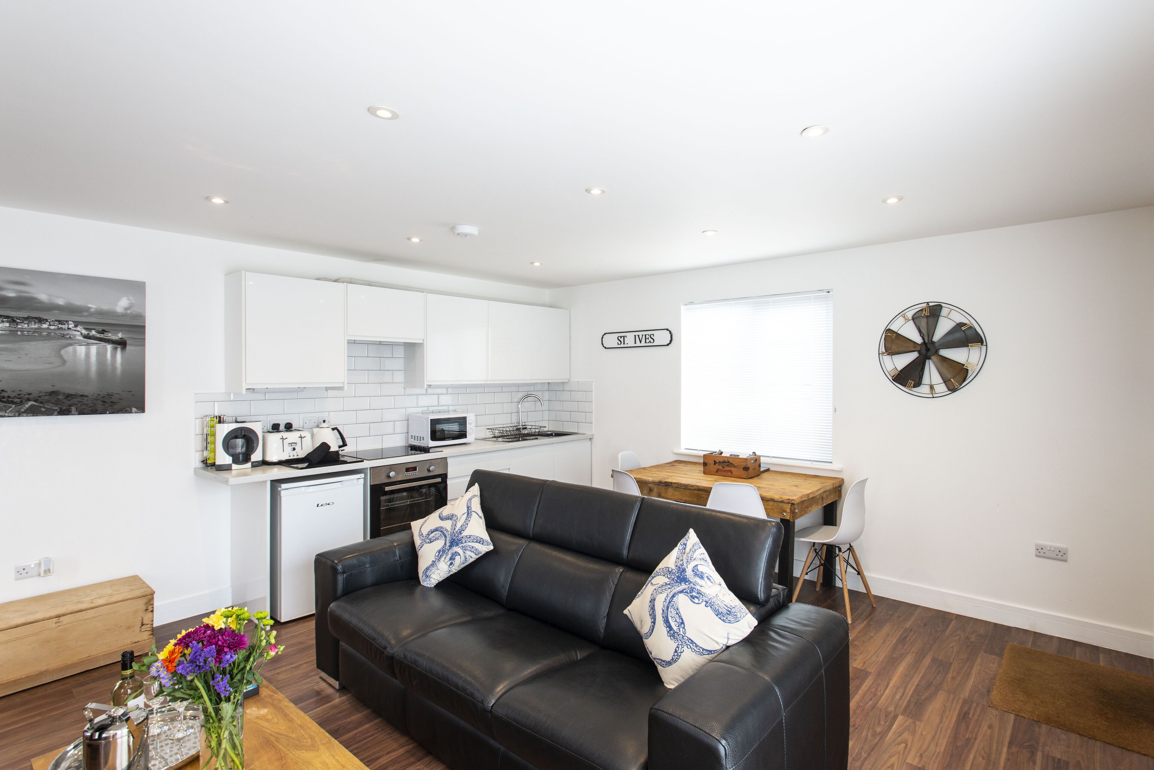 A modern living room with a black leather sofa, a wooden coffee table, and a dining area with a wooden table and white chairs. A kitchen with white cabinets and a microwave is visible in the background.