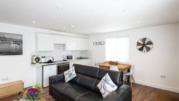 A modern living room with a black leather sofa, a wooden coffee table, and a dining area with a wooden table and white chairs. A kitchen with white cabinets and a microwave is visible in the background.