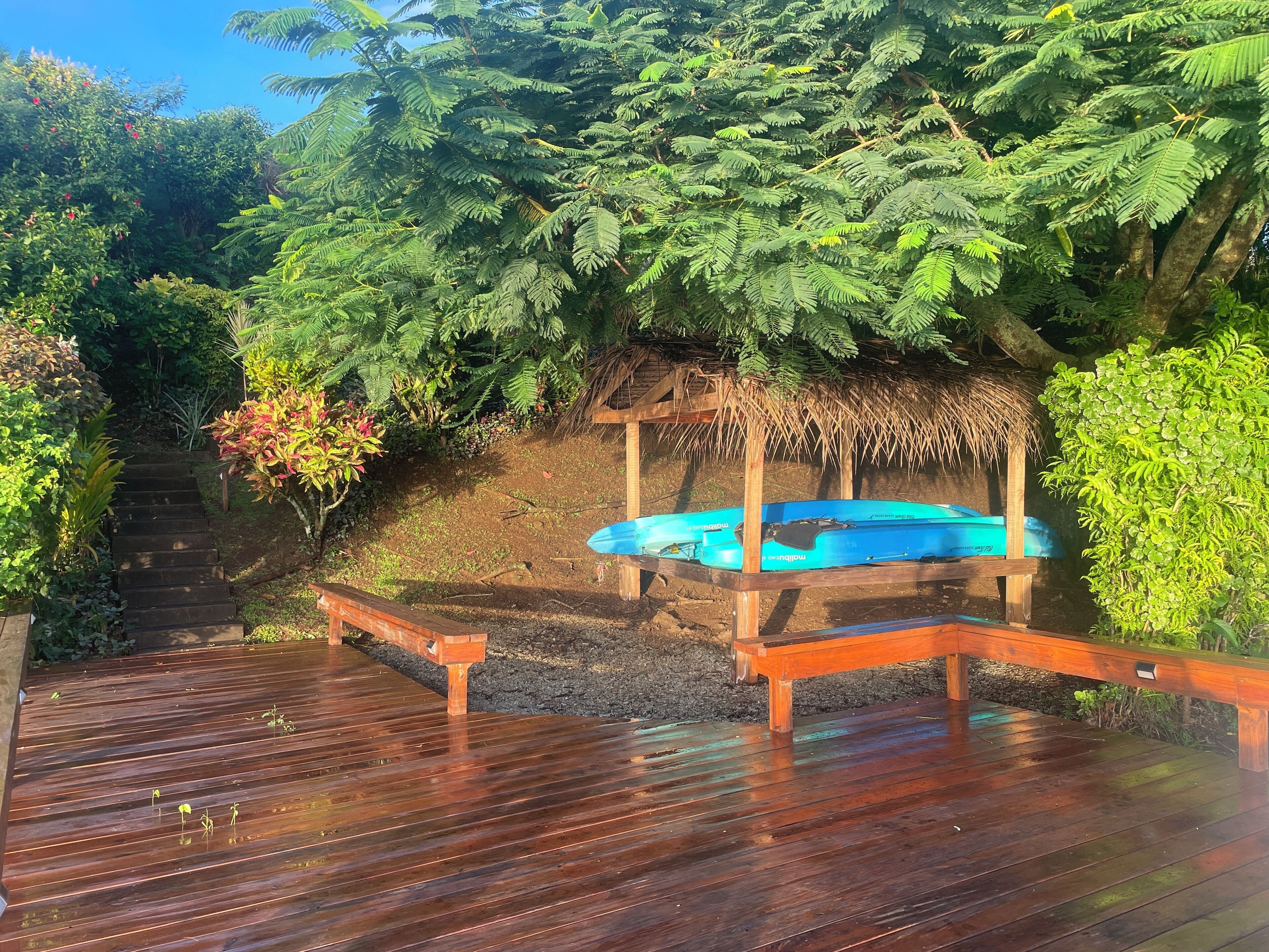 Une terrasse en bois avec un kayak bleu, entourée d’une végétation luxuriante et d’une structure à toit de chaume.