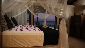 Ein Bett mit weißem Baldachin, einer schwarzen Tagesdecke und Blick auf das Meer durch große Fenster.