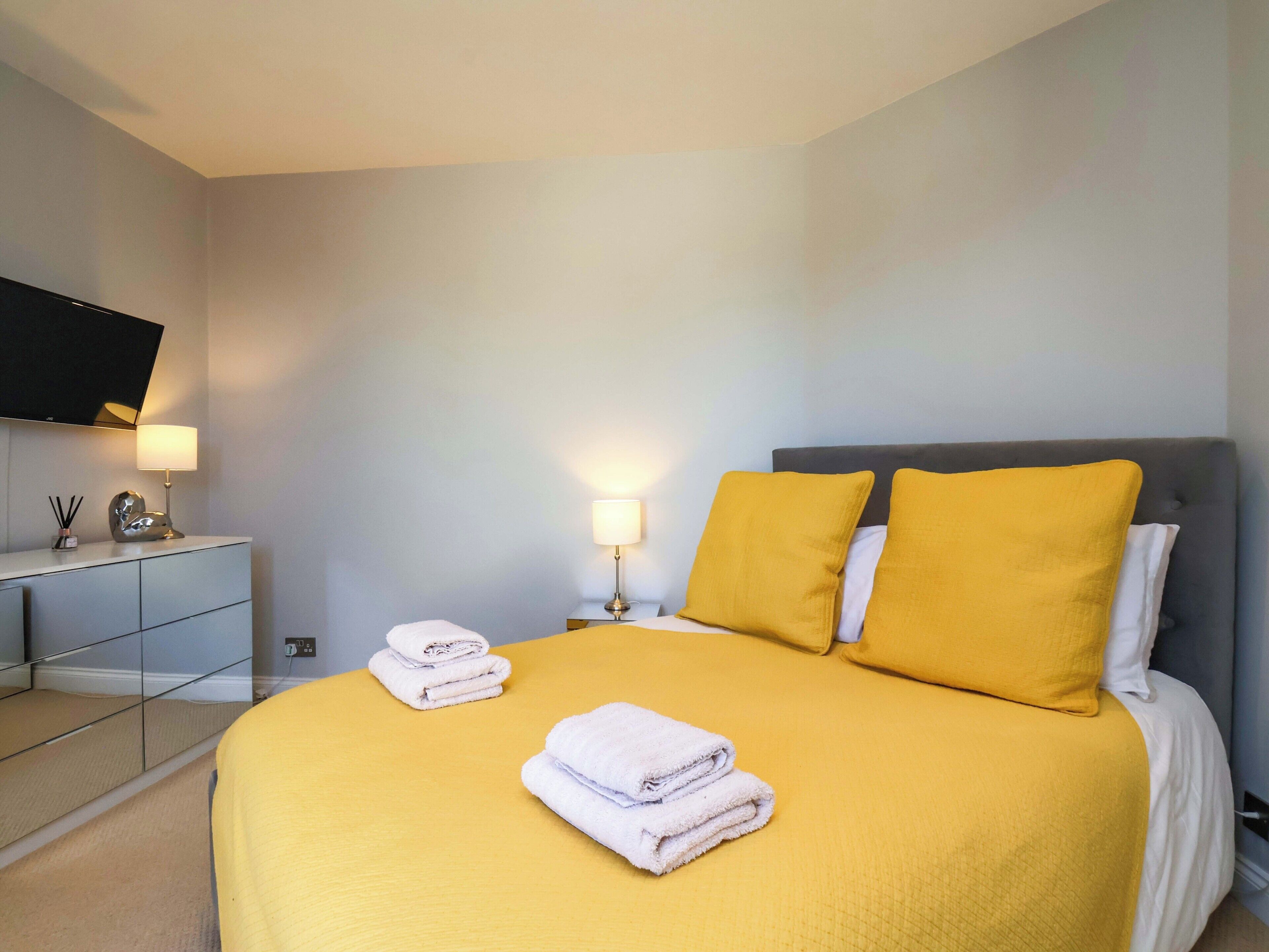 Une chambre d’hôtel avec un lit, deux lampes de chevet, un téléviseur à écran plat fixé au mur et une petite table sur laquelle se trouvent une lampe et quelques objets décoratifs.