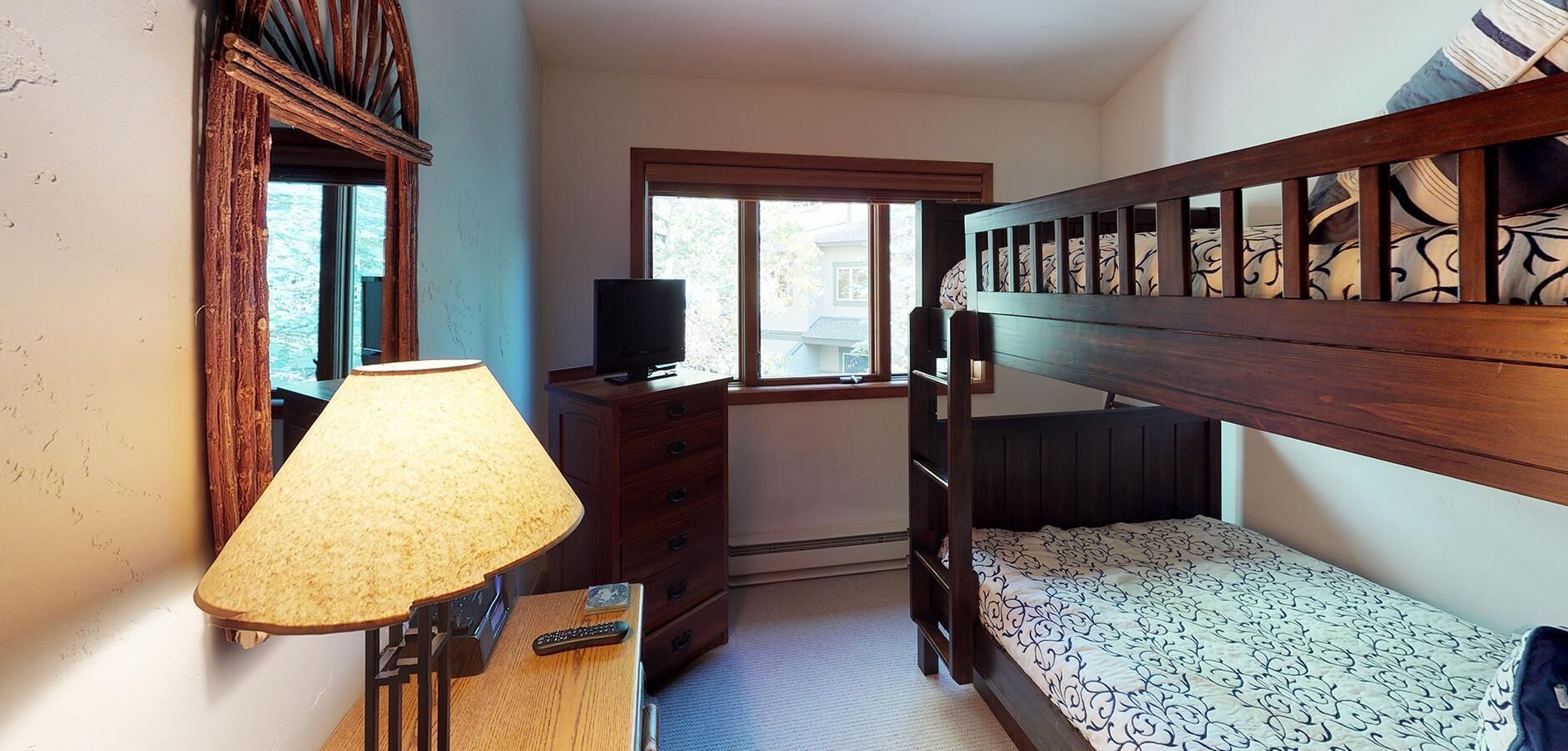 A bedroom with a bunk bed, a desk with a lamp, a TV, and a window.