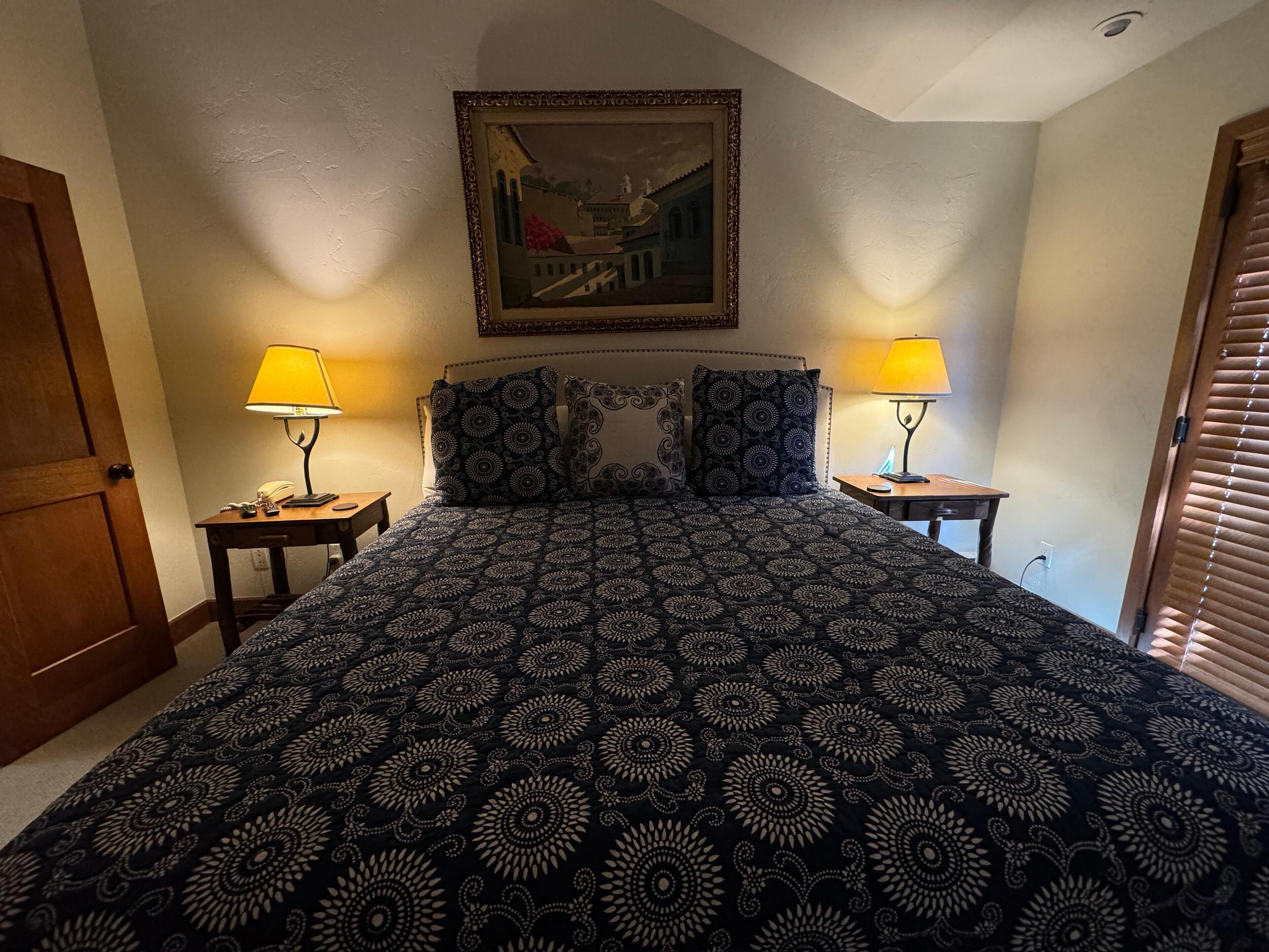Ein Schlafzimmer mit einem gemusterten Bett, zwei Nachttischlampen, einem Bild an der Wand und einer Tür links.