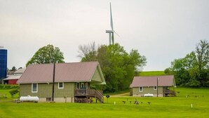 Paesaggio rurale con due edifici, uno con portico e un silo, e una turbina eolica sullo sfondo.