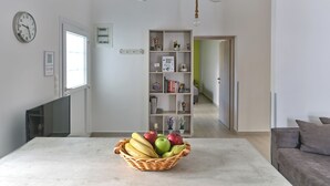 Una sala de estar moderna con una mesa grande, un televisor y una cesta de frutas. Hay tres lámparas colgantes y una estantería al fondo.