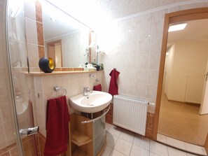 Une salle de bain avec un lavabo blanc, un miroir et un porte-serviettes. Il y a un espace douche avec une porte en verre et une serviette rouge accrochée au mur.