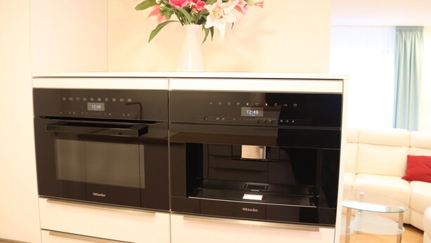 Una cocina con un horno y microondas Miele negros, un jarrón blanco con flores en la pared y un sofá blanco con una almohada roja al fondo.