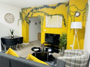 A living room with a yellow accent wall, a black TV stand, a plaid sofa, and a dining table with chairs.