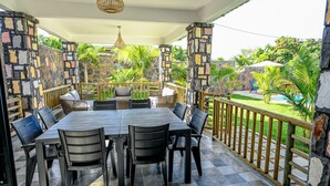 Un patio couvert avec un mur en pierre, une table à manger avec des chaises et une vue sur une piscine et la verdure.