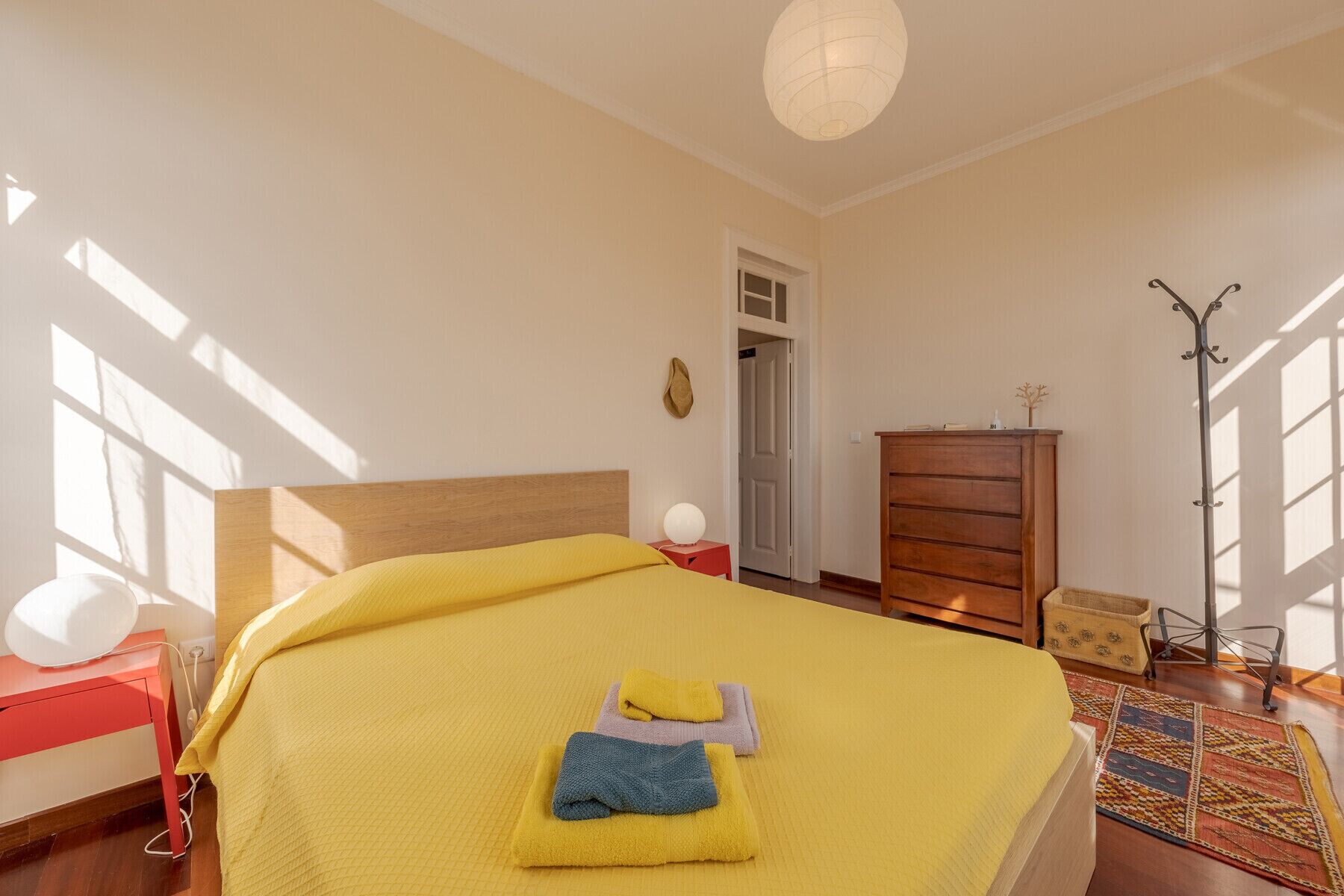A bedroom with a bed, a wooden dresser, a red nightstand, a lamp, and a door leading to another room.