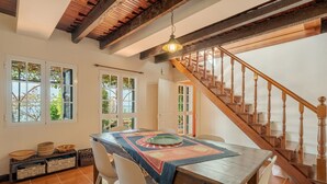 Un coin repas avec une table en bois, des chaises blanches et un chemin de table à motifs. Un escalier avec des rampes en bois mène à l’étage supérieur.