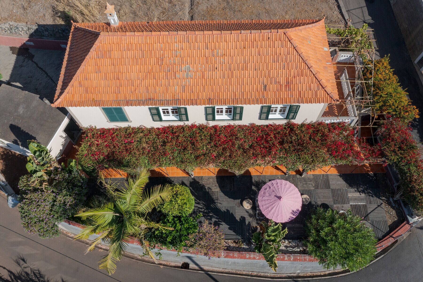 Une vue aérienne d’un immeuble à deux étages, doté d’un toit recouvert de tuiles, d’un balcon orné de fleurs rouges et d’une terrasse en bois avec un parasol rose.