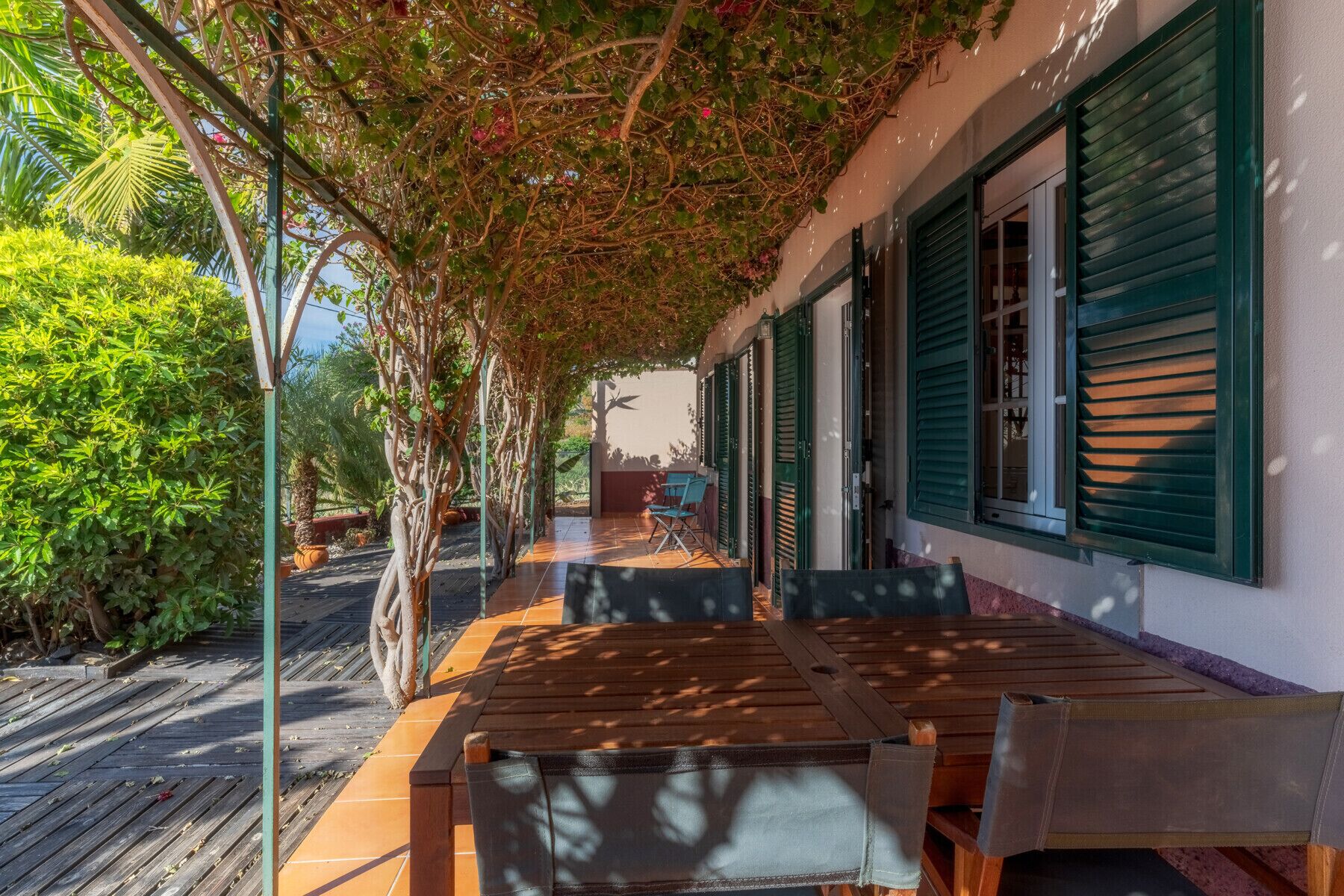 Un espacio al aire libre cubierto con mesas y sillas de madera, una terraza de madera y vistas a un edificio con contraventanas verdes.