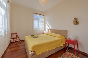 A bedroom with a wooden bed, a red nightstand, a white lamp, and a window with curtains. - Villa Flora (Calheta)